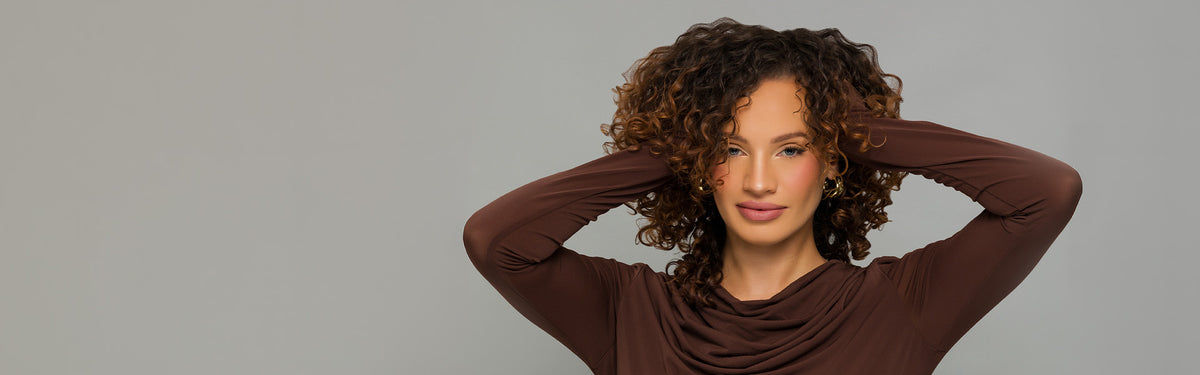 A woman with curly brown hair wearing a brown long-sleeve top, posing with her hands behind her head against a solid gray background.